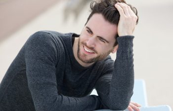 A man in a dark sweater leans on an outdoor bench, smiling and resting his head on one hand.