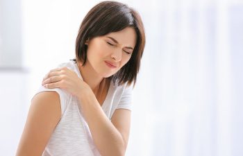 A woman in a white shirt holds her shoulder with a pained expression.